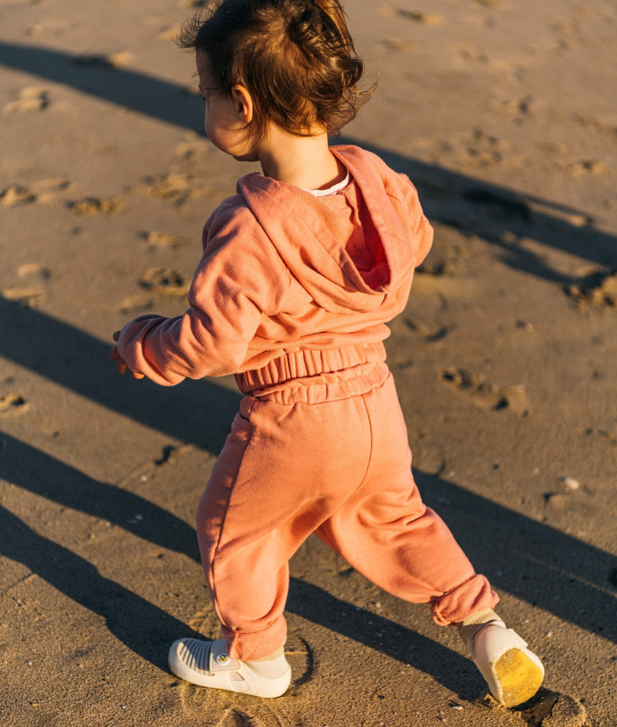 A baby in an orange tracksuit, walking on the beach, wearing Pediatrician-approved jbrds baby walking shoes.