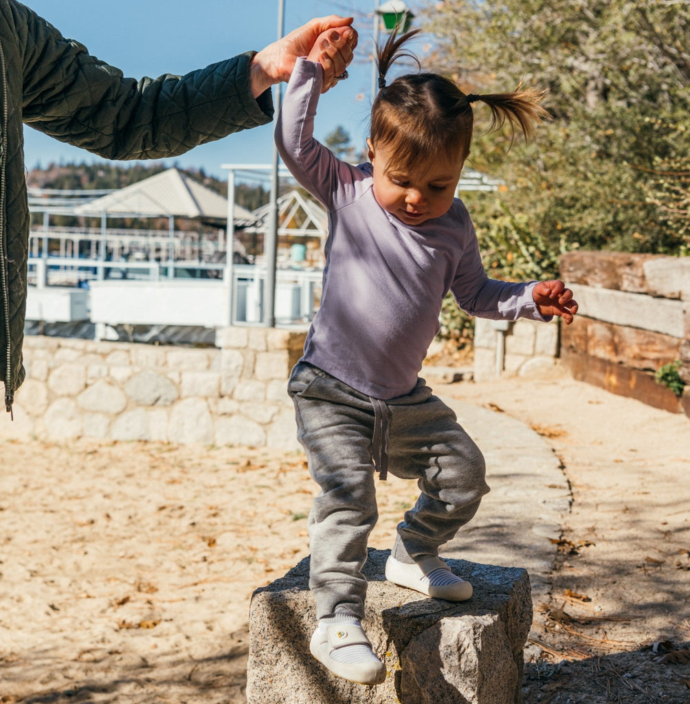 A child balancing on a stone block outdoors wearing jbrds first walker shoes, accompanied by an adult.