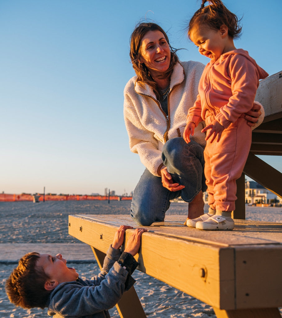 A toddler wearing jbrds shoes and playing with another toddler and an adult woman on the beach.